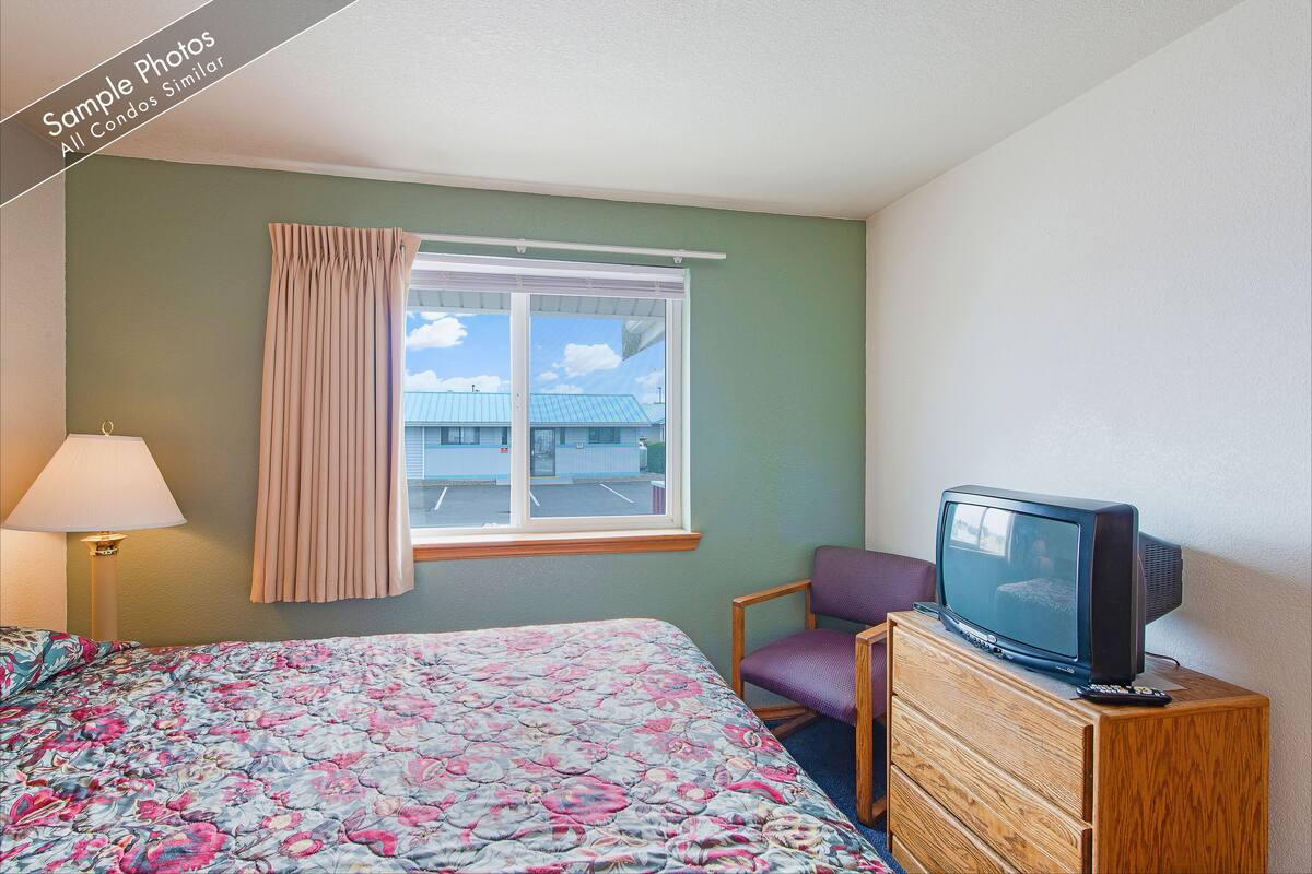 A hotel room with a bed, a television on a wooden dresser, a chair, a lamp, and a window with curtains.