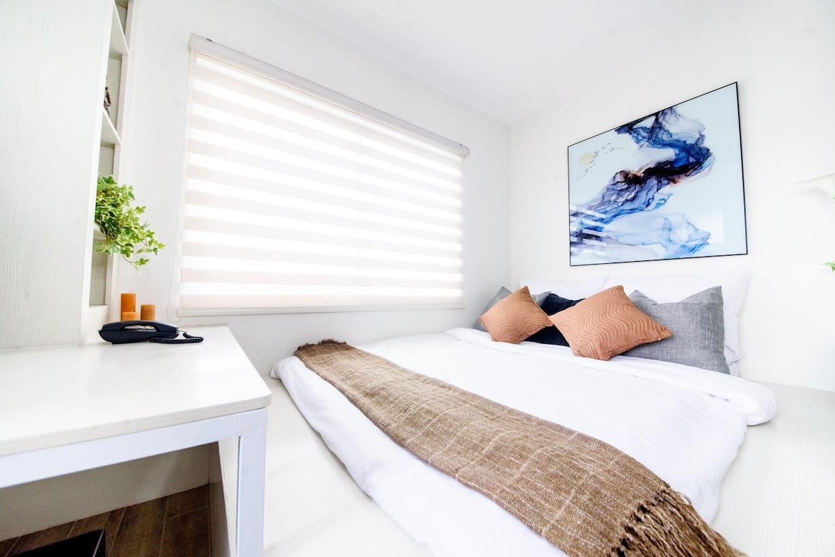 A bedroom with a bed, white walls, a window with blinds, a bedside table with a phone and a plant, and a painting on the wall.