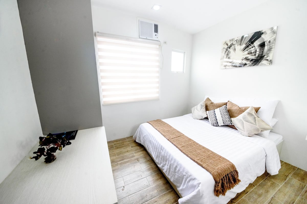 A bedroom with a bed, a window with blinds, a wall-mounted air conditioner, and a small wall art piece.