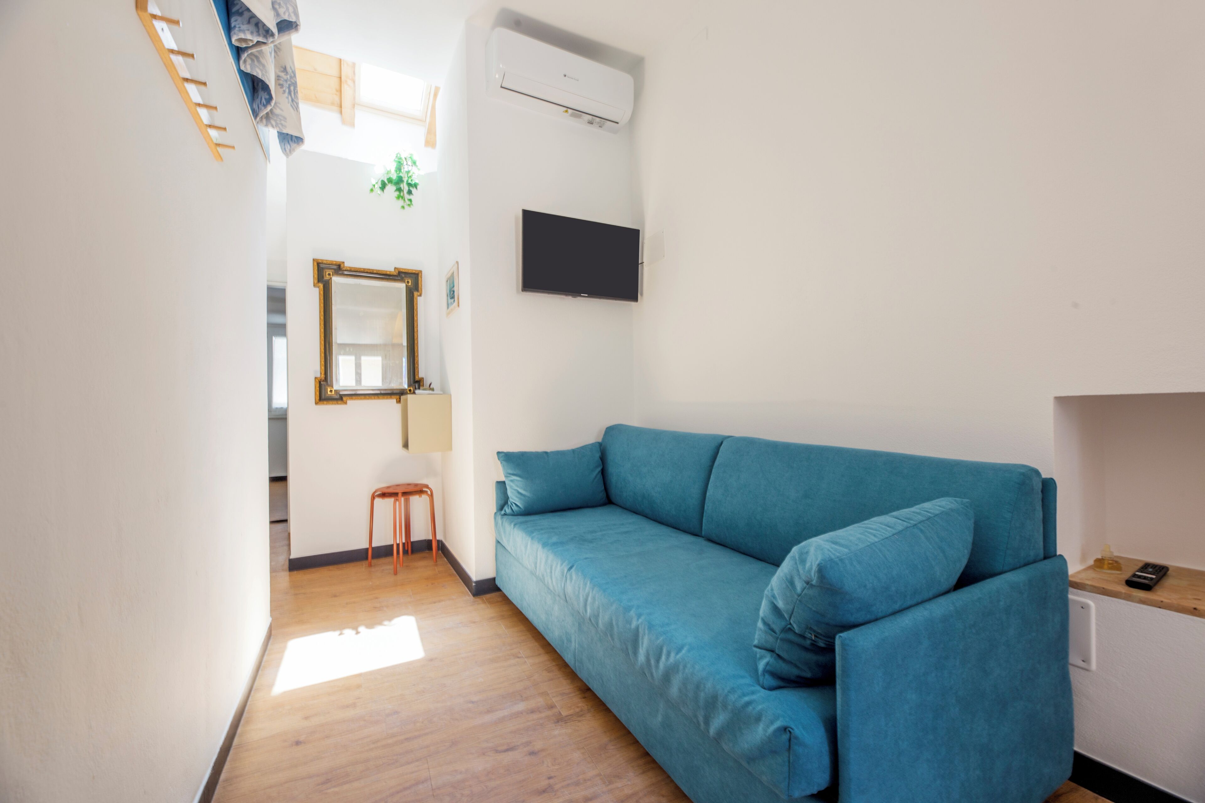 Une pièce avec un canapé bleu, un plancher en bois, un téléviseur fixé au mur et un petit tabouret.