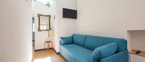 Une pièce avec un canapé bleu, un plancher en bois, un téléviseur fixé au mur et un petit tabouret.