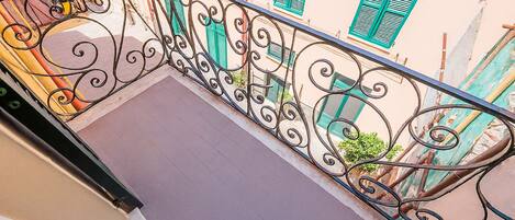 Un balcón con barandillas de hierro forjado negro ornamentado, piso de baldosas y edificios con contraventanas verdes al fondo.