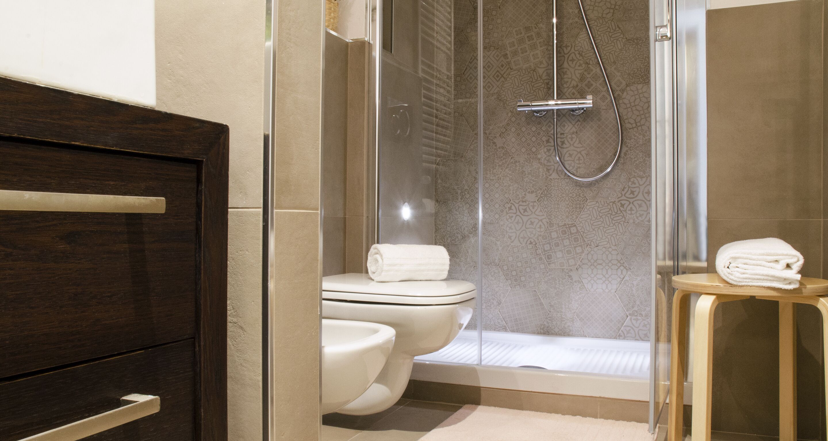 A modern bathroom with a glass-enclosed shower, a toilet, and a wooden stool with a towel.