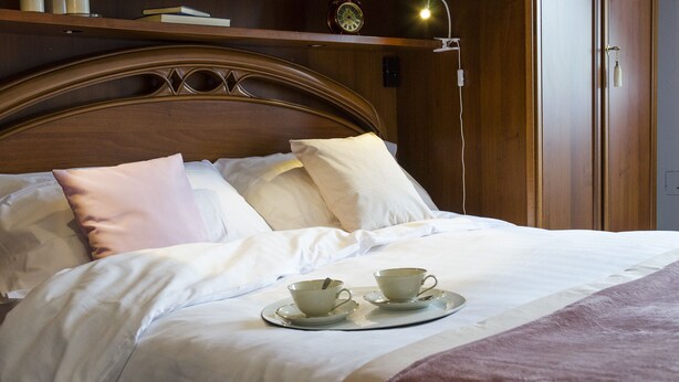 A neatly made bed with white linens, two cups and saucers on a tray, a clock, and a bedside lamp on a wooden nightstand. - JOIVY Little House (Como)