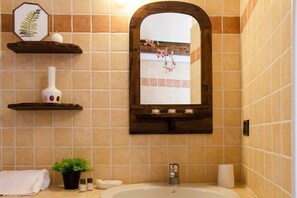 A bathroom with a bathtub, a wooden shelf with decorative items, a potted plant, and a mirror with a floral arrangement. - JOIVY Attic on the Lake (Argegno)