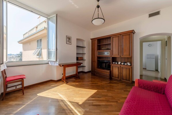 A spacious living room with a wooden floor, a red sofa, a wooden table, and a built-in wooden cabinet. A window provides a view of the outside.