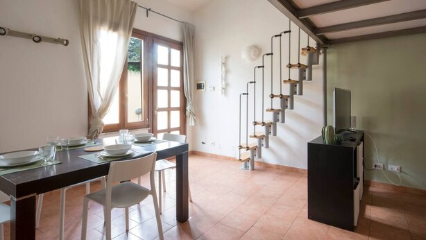 A dining area with a table set for four, a staircase with a unique design, and a television on a stand. - JOIVY Green Terraced House (Pisa)