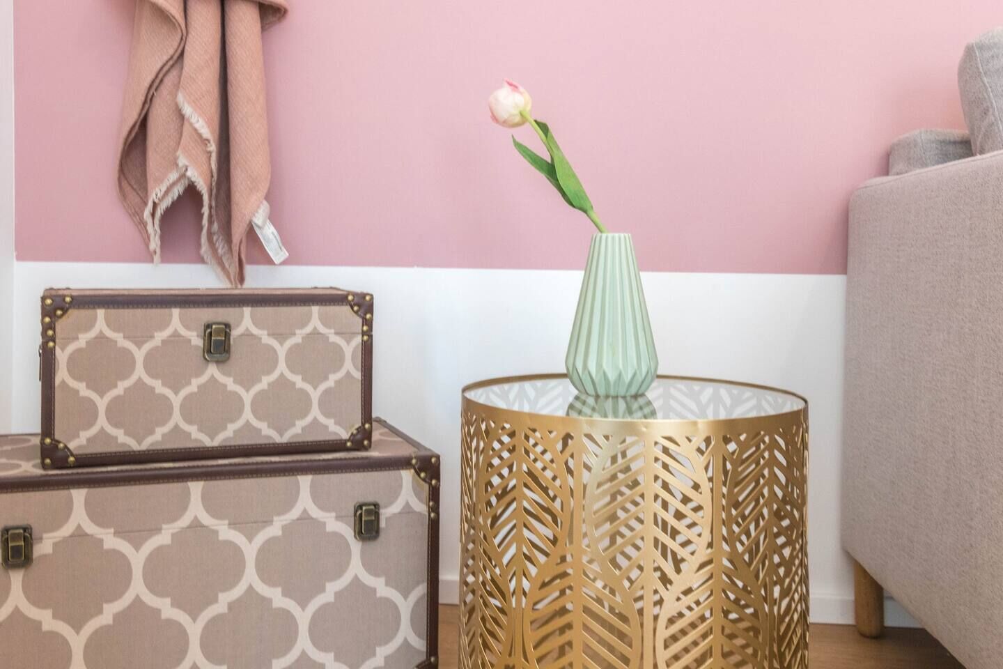 Una habitación con pared rosa, sofá beige, maleta estampada, jarra de vidrio con una flor y una mesita de noche de madera decorativa.