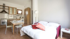 A modern kitchen with a dining area, a bed with red pillows, and a bedside table with a glass object. - JOIVY Red Terraced Apartment (Pisa)