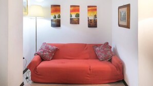 A red sofa with floral cushions in a room with a white wall and three vertical paintings depicting landscapes. - JOIVY Villa Il Glicine sul Mare (Pietrasanta)
