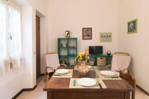 A dining area with a wooden table set for four, surrounded by chairs. A green shelving unit with decorative items and a television is in the background. - JOIVY Villa Il Glicine sul Mare (Pietrasanta)