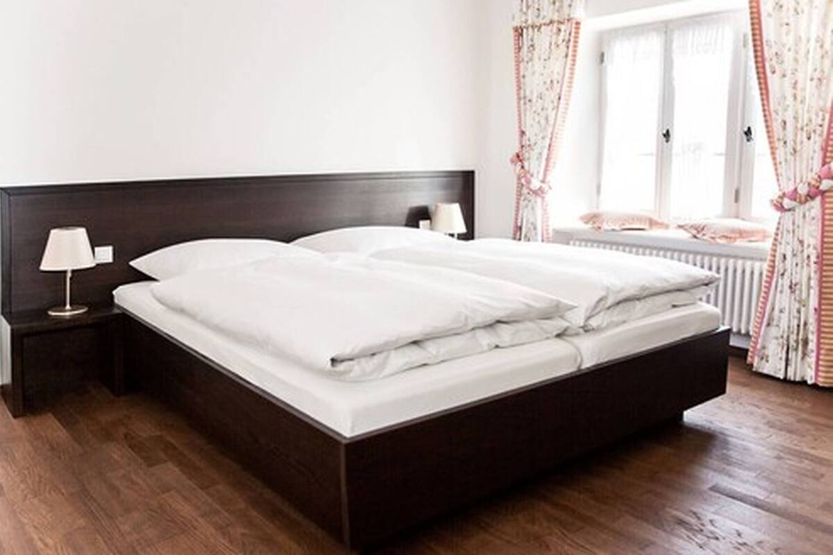 Un lit bien fait dans une chambre avec une tête de lit en bois foncé, une table de chevet avec une lampe et une fenêtre ornée de rideaux à motifs floraux.