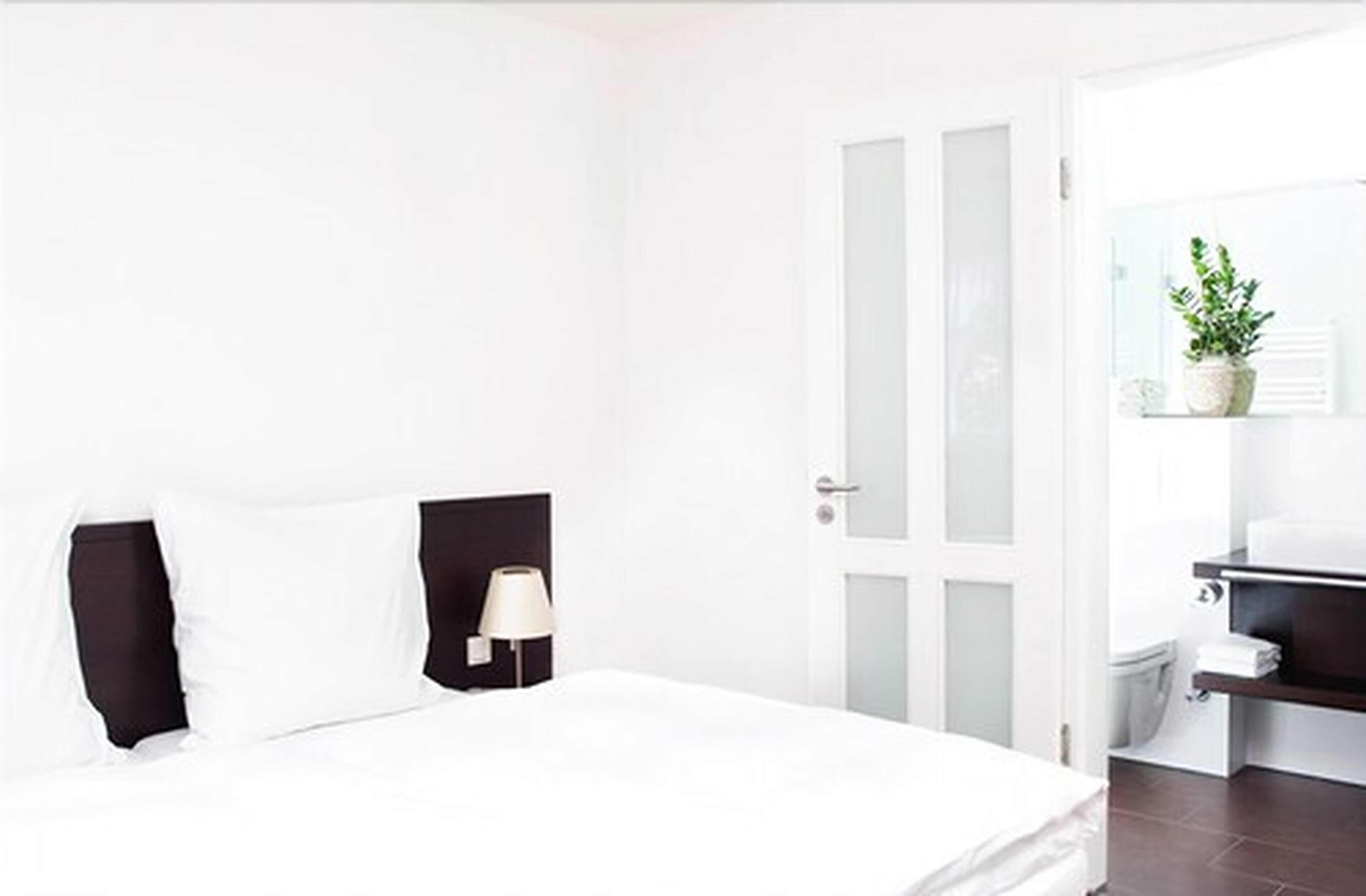 A hotel room with a bed, a nightstand, a lamp, a bathroom with a toilet, and a potted plant.