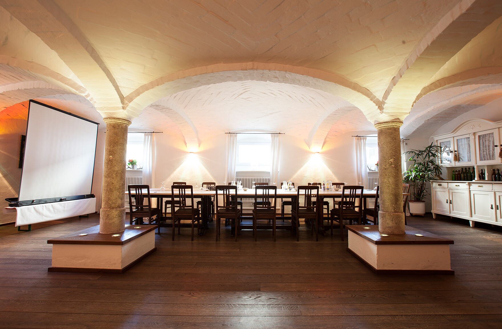 A spacious dining area with a high ceiling and exposed brick walls, featuring a long table with chairs and a whiteboard on the left.