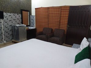A bedroom with a bed, a chair, a wardrobe, a mirror, and a small table. - Hotel Sweet Inn (Lahore)