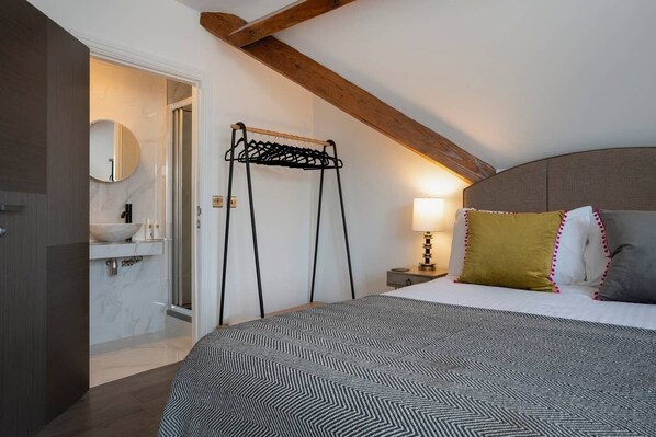 Een slaapkamer met een bed, nachtkastje, lamp, kledingrek en een badkamer met wastafel en spiegel.
