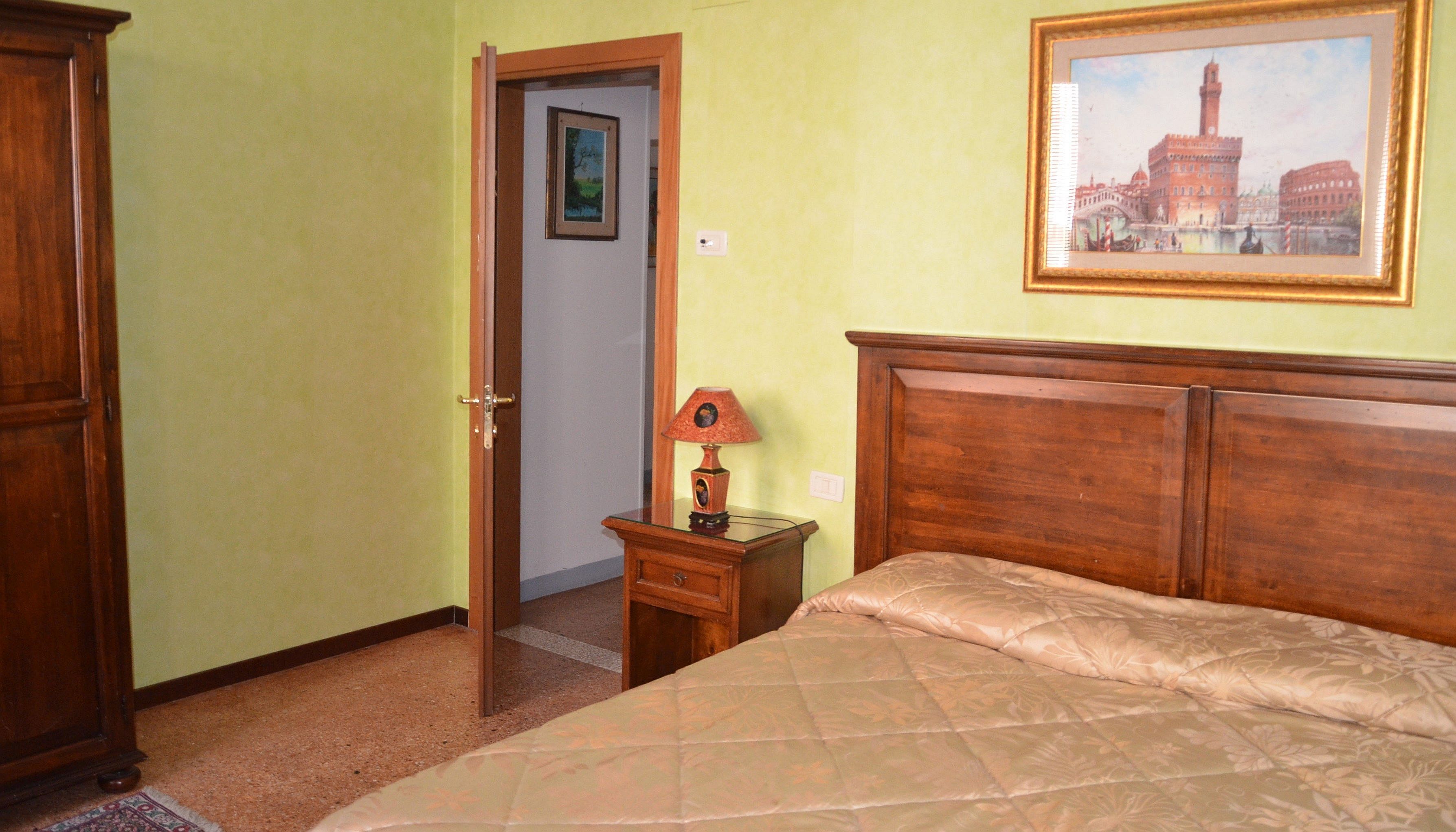 A bedroom with a wooden bed, a nightstand with a lamp, a door leading to another room, and a framed picture on the wall.