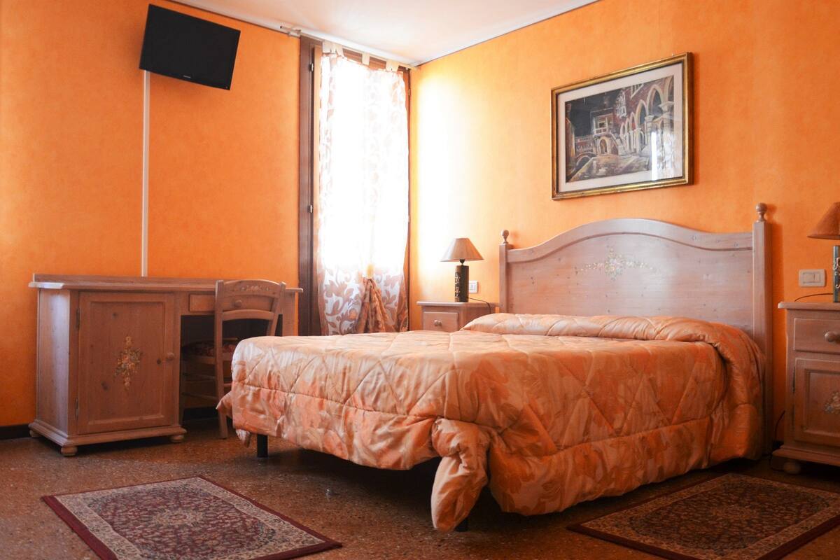 Ein Schlafzimmer mit einem Bett, einem Schreibtisch, einem Sessel, einem Nachttisch, einer Lampe, einem Fernseher und einem Bild an der Wand.