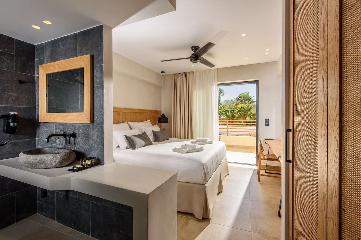 A modern hotel room with a stone sink, a large bed, a ceiling fan, and a view of the outdoors.