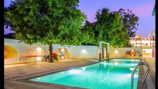 An outdoor swimming pool with a tiled deck, surrounded by trees and a wall with murals. - Dasmaya Resort (Udaipur)