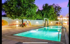 Una piscina exterior con borde de baldosas, rodeada de árboles y un muro con murales.