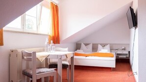 A small, well-lit room with a bed, a table with chairs, a radiator, and a television mounted on the wall. - Hotel Garni (Isny im Allgaeu)