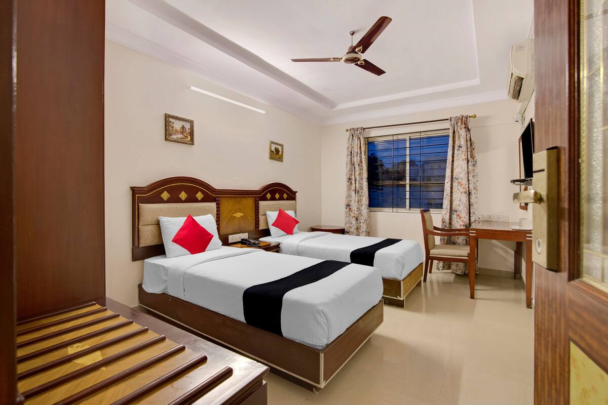 Une chambre d’hôtel avec deux lits, un bureau, une chaise, un ventilateur de plafond et une fenêtre avec des rideaux.