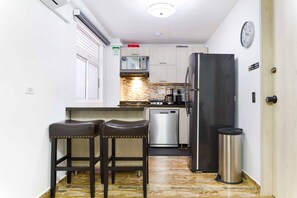 A compact kitchen with a refrigerator, microwave, and dishwasher. There are two stools and a trash can.