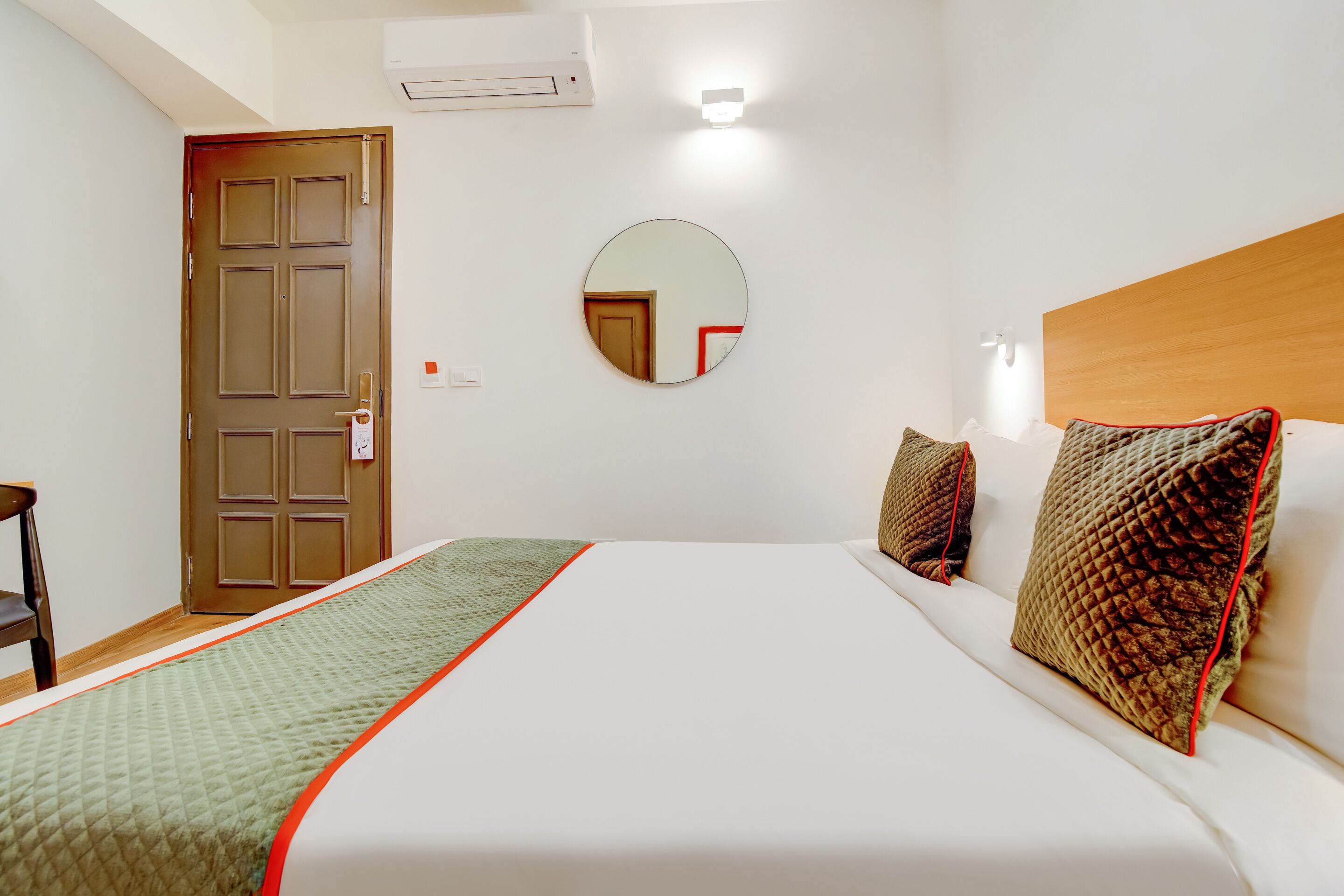 A hotel room with a bed, a round mirror, a wooden door, and a wall-mounted air conditioner.