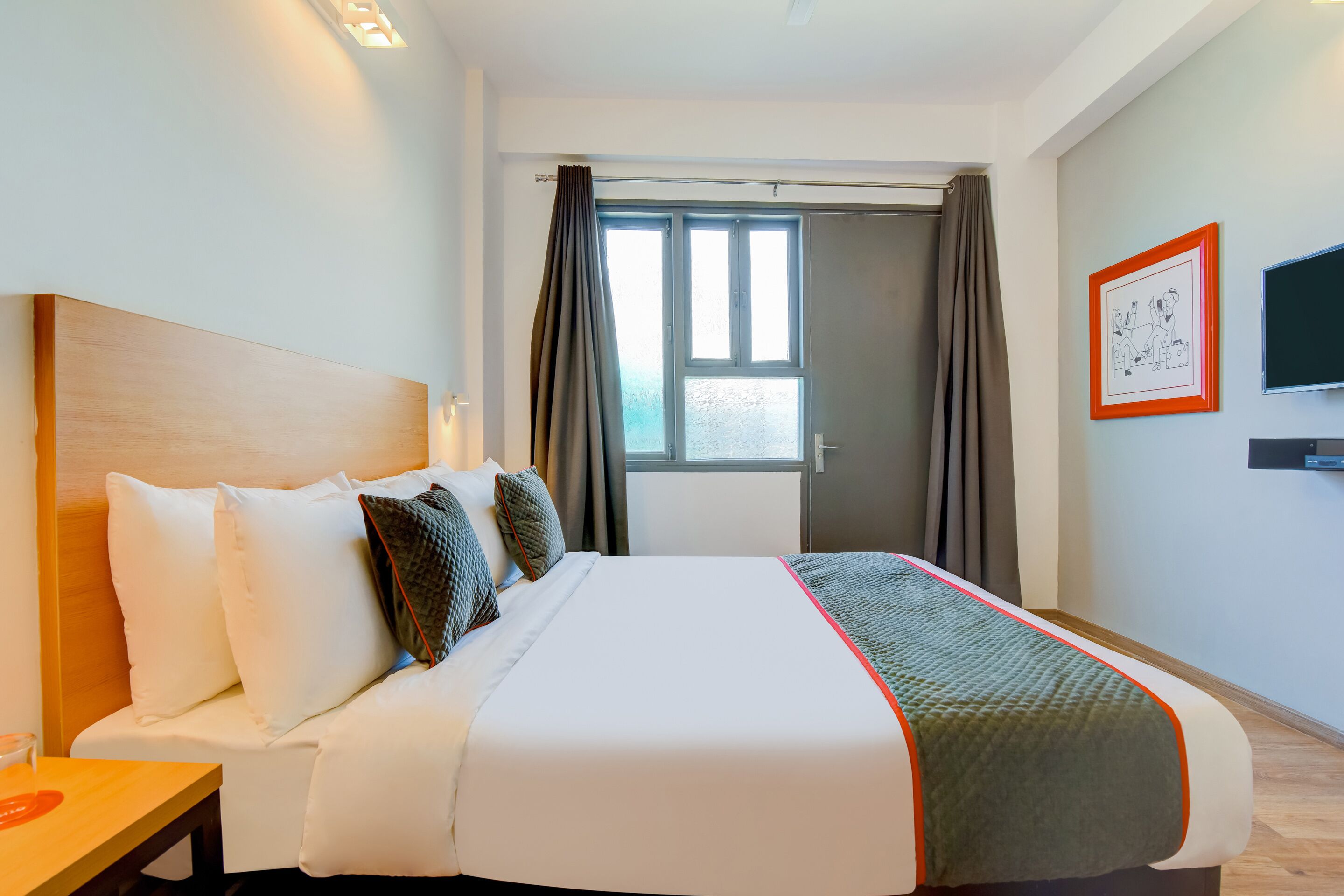 A hotel room with a large bed, a bedside table, a TV mounted on the wall, and a window with curtains.