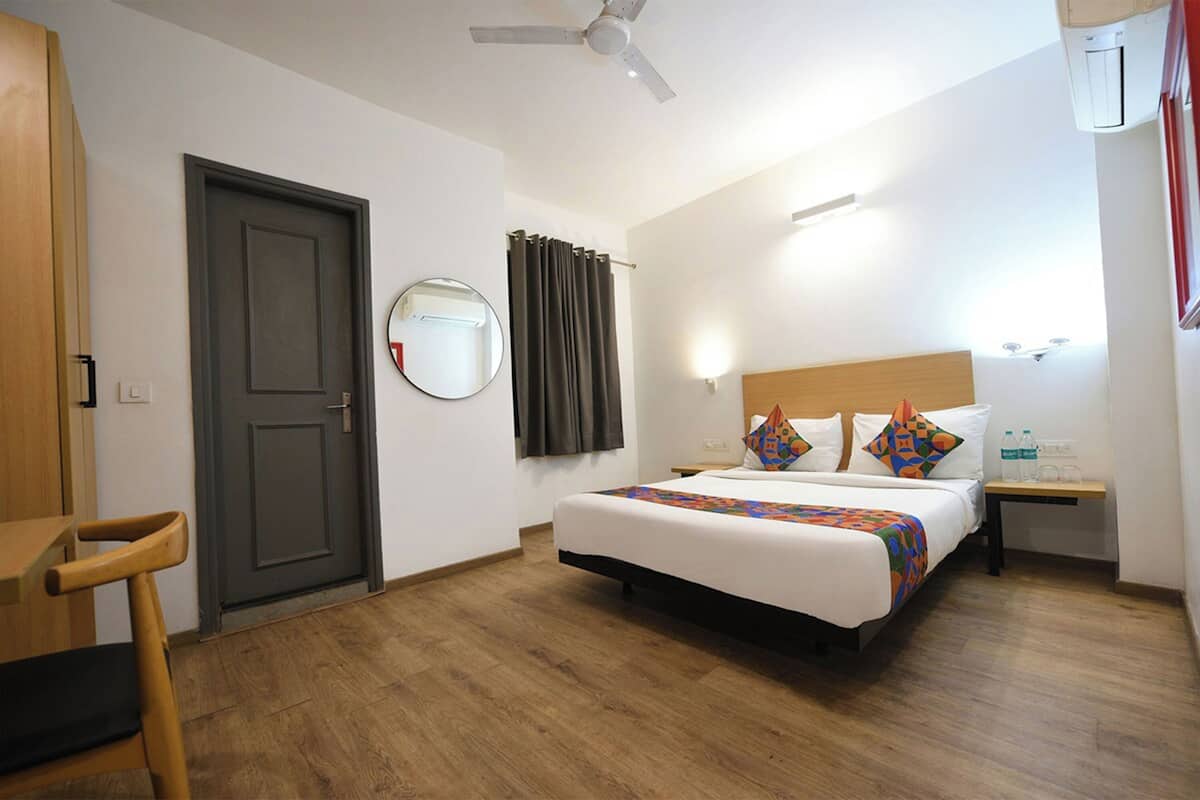 Habitación de hotel con cama, escritorio, silla, ventilador de techo, espejo, mesita de noche y unidad de aire acondicionado.
