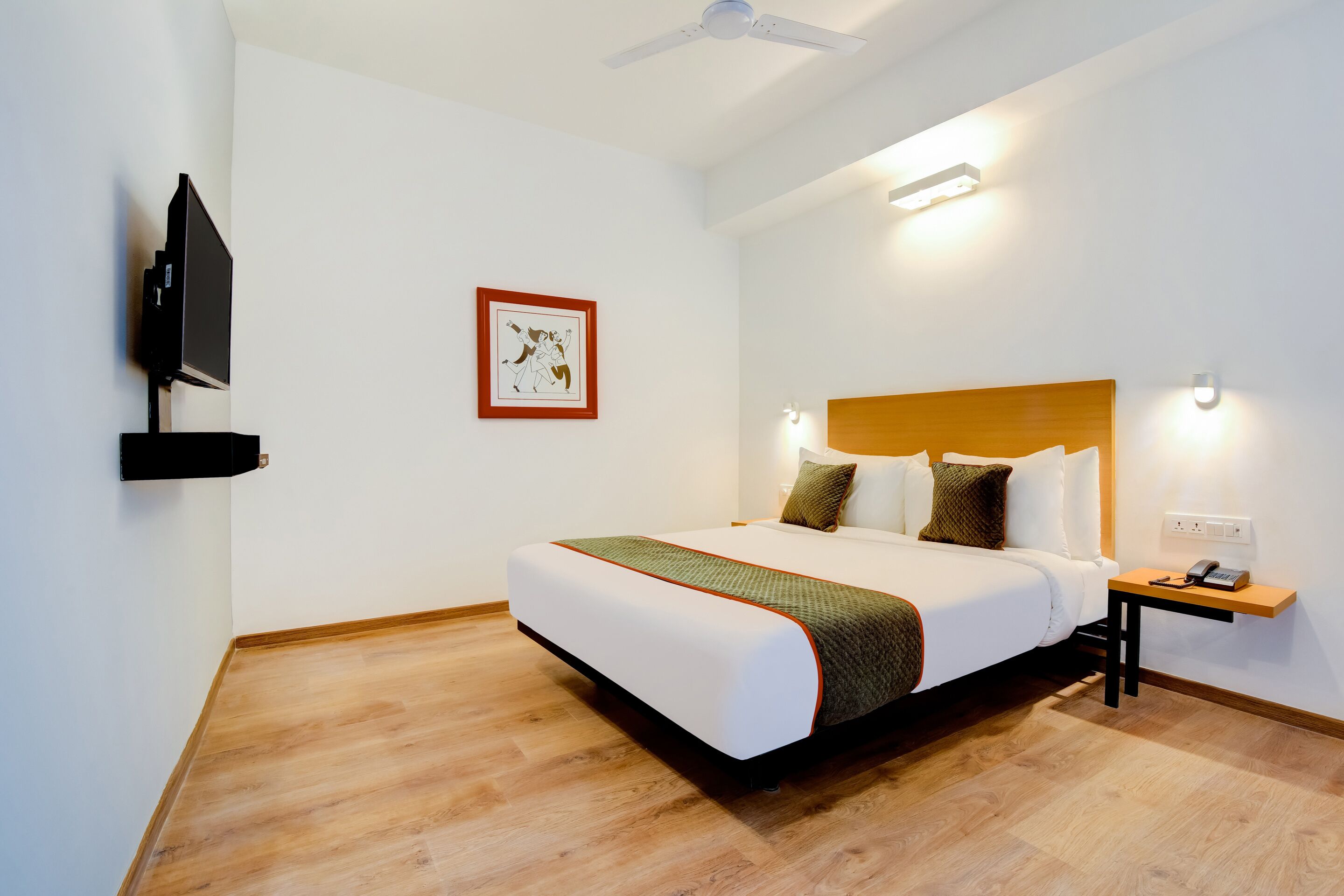 A hotel room with a bed, a bedside table, a wall-mounted TV, and a framed picture on the wall.