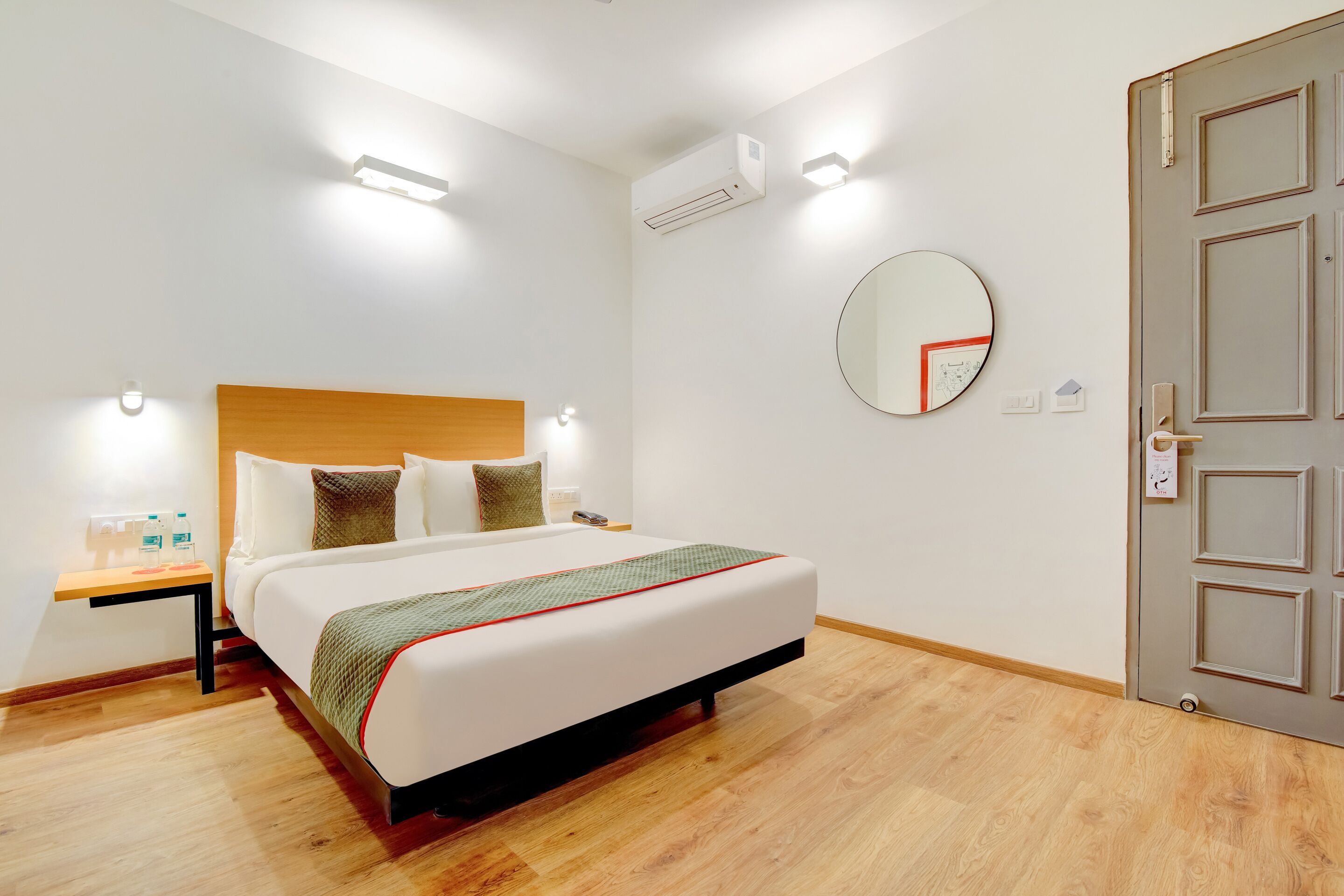Quarto de hotel com cama grande, mesas de cabeceira, espelho redondo e porta com puxador.