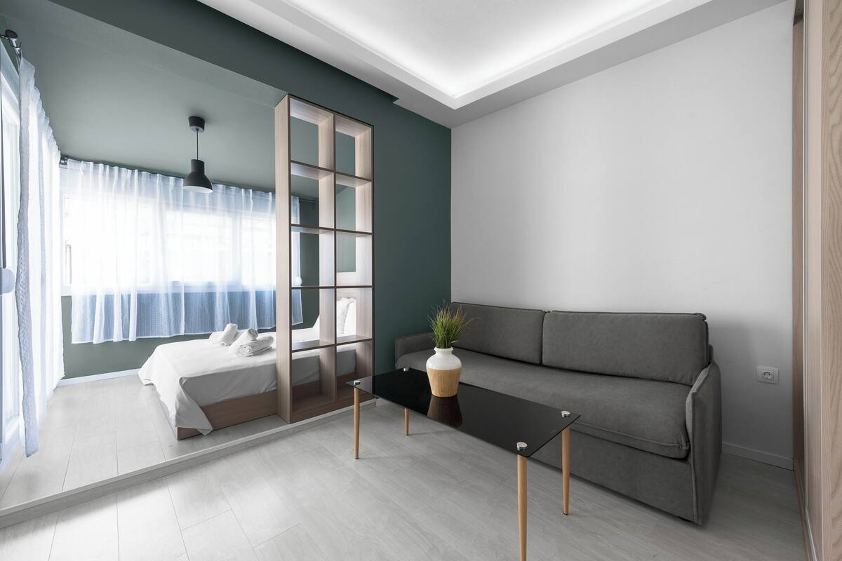 A modern room with a bed, a grey sofa, a black coffee table, and a large window with sheer curtains.