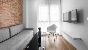 A modern living room with a brick wall, a gray sofa, a white chair, a black coffee table, and a flat-screen TV mounted on the wall. - DeniZen Boutique Apartments, Nilie Hospitality MGMT (Thessaloniki)