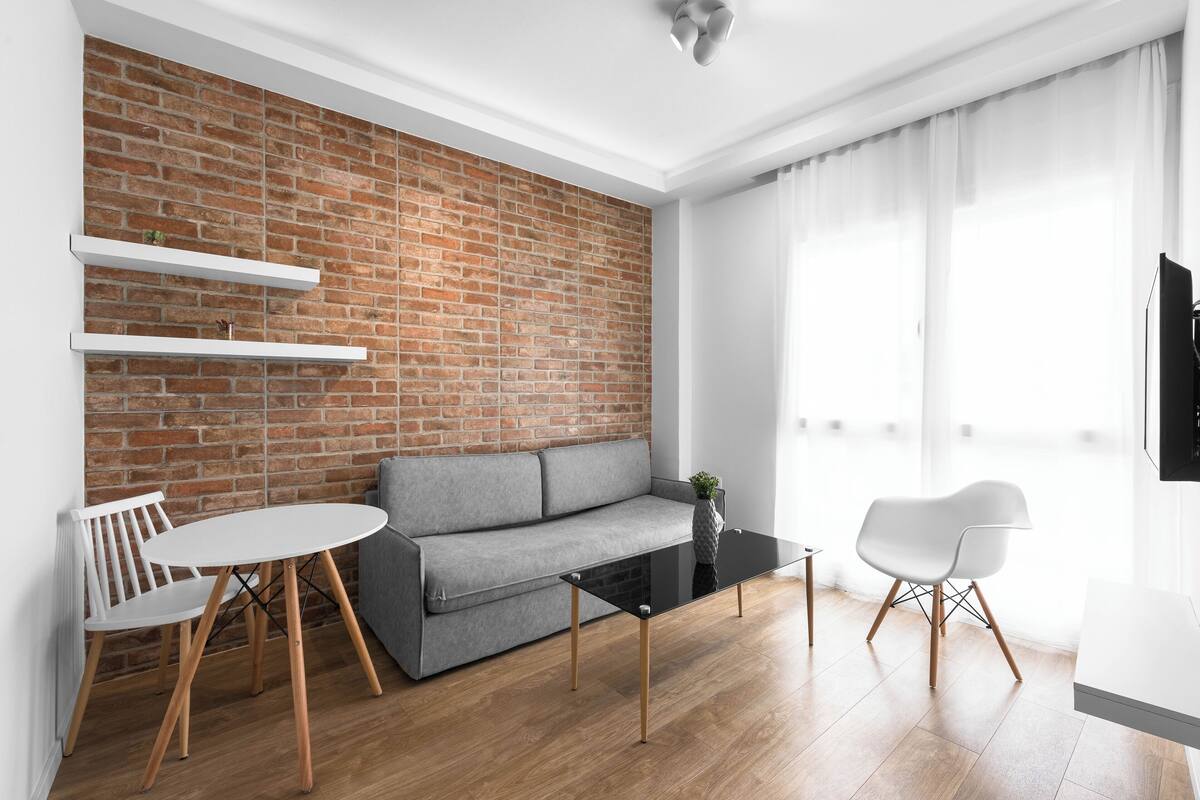 A modern living room with a brick wall, a grey sofa, a round glass coffee table, white chairs, and a flat-screen TV.