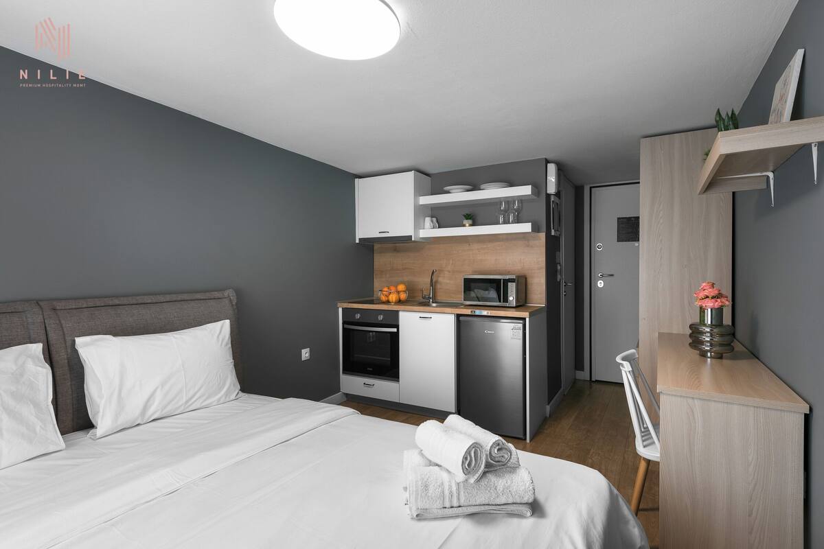 A modern hotel room with a bed, a kitchenette, and a desk.