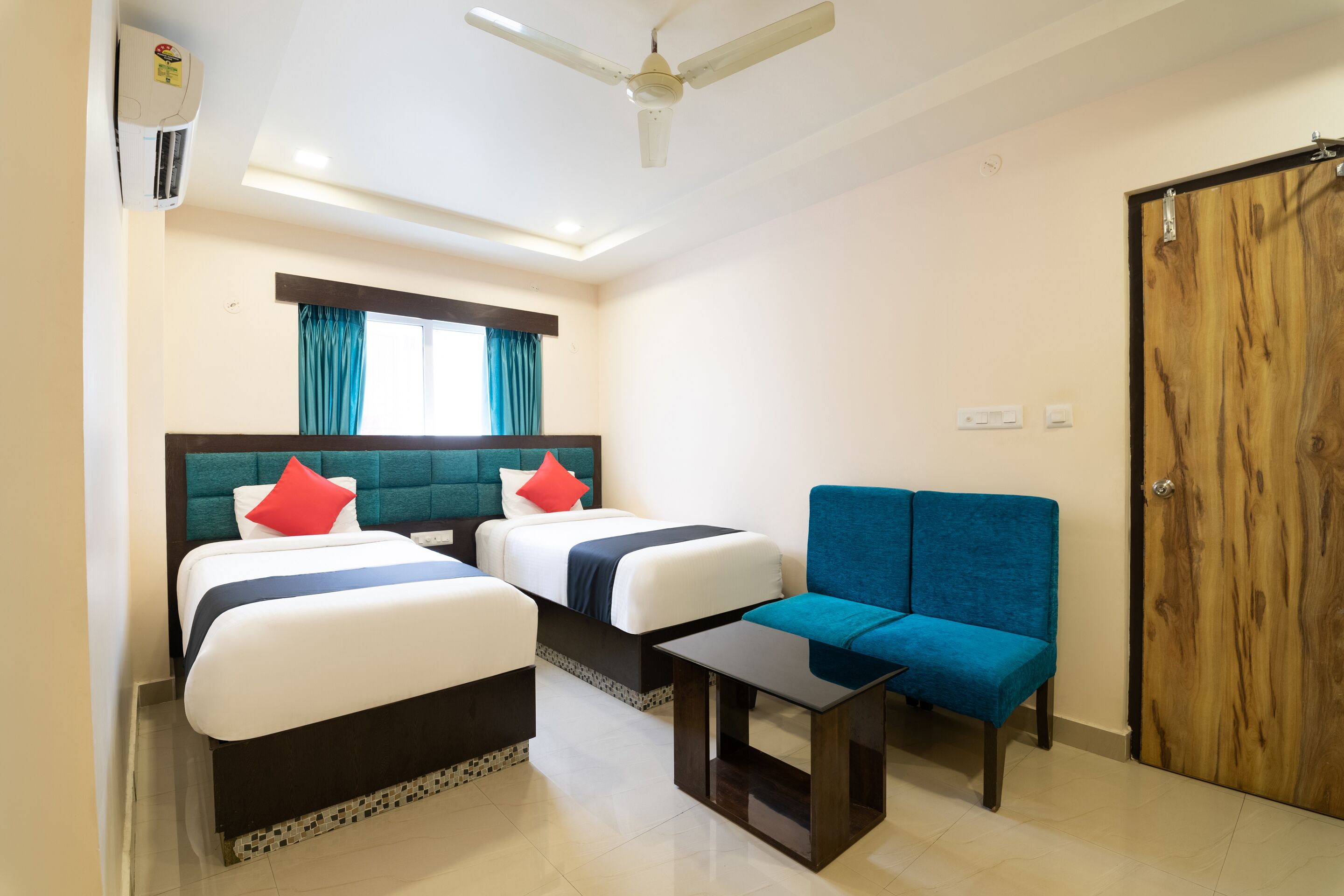 A hotel room with two beds, a blue sofa, a small table, a ceiling fan, and a wooden door.