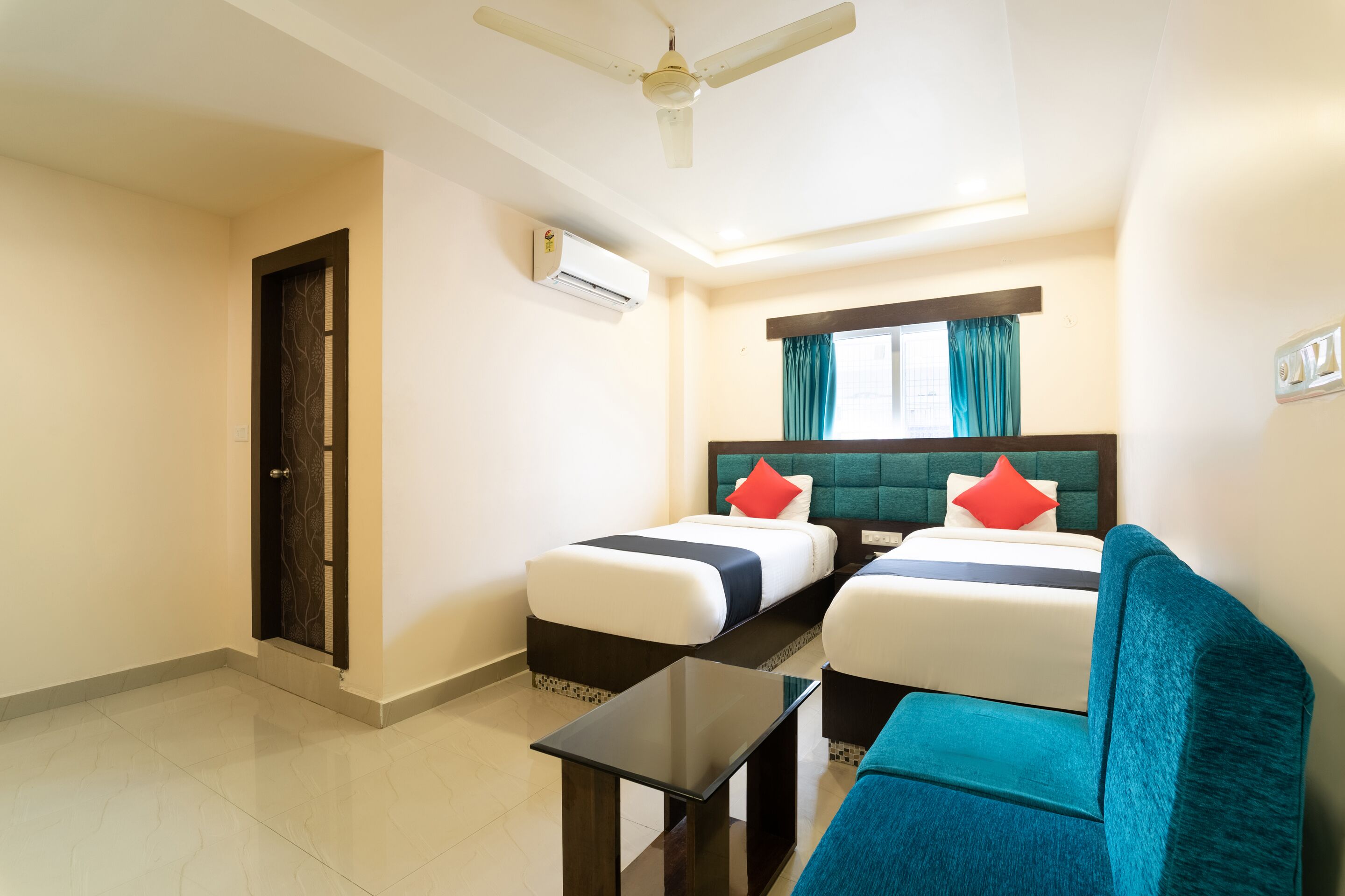 A hotel room with two beds, a ceiling fan, an air conditioning unit, a window with curtains, and a small table.