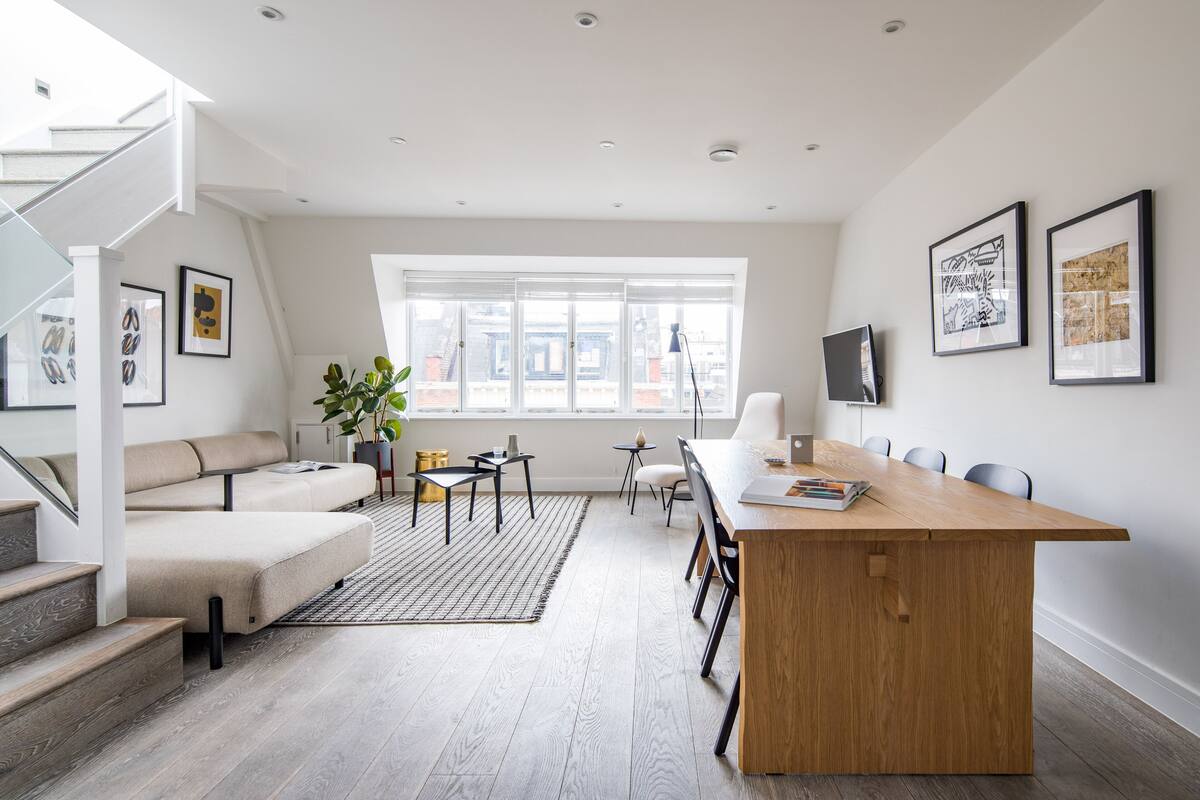 A modern living room with a wooden dining table, a sofa, and a staircase.