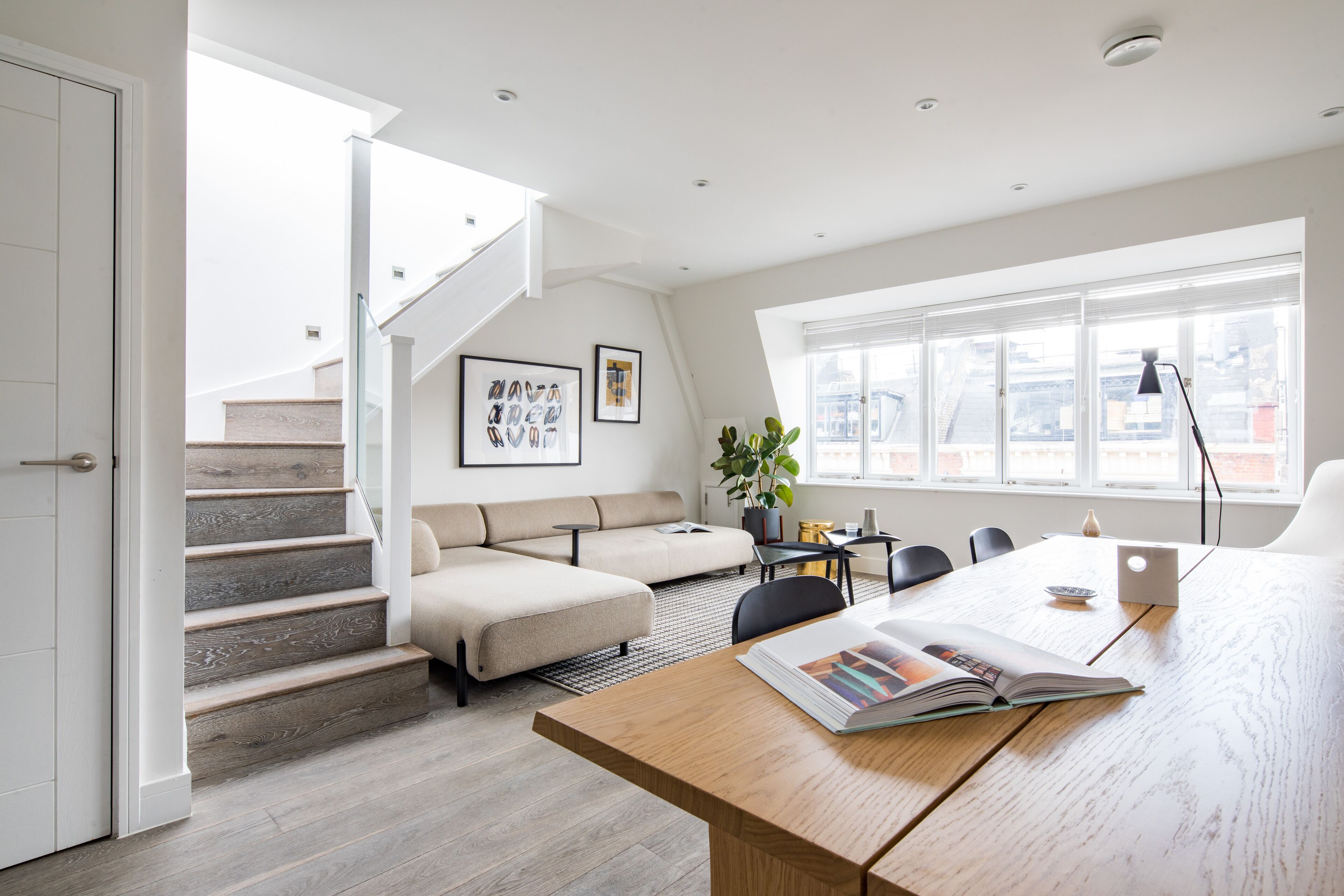 Una sala de estar moderna con una escalera, un sofá, una mesa de comedor y un ventanal grande.