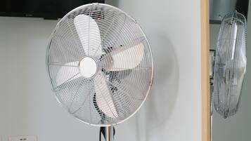A standing fan with a metallic grill and a central hub, positioned against a plain wall with a power outlet visible.