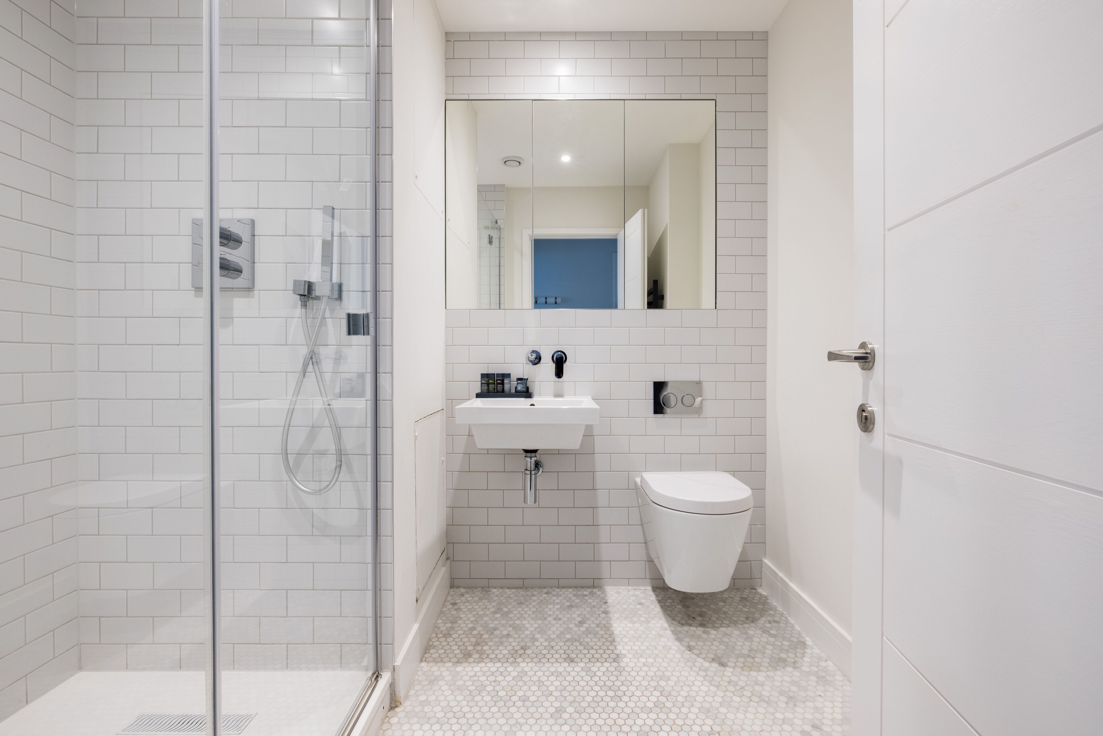Un baño moderno con paredes de azulejos blancos, cabina de ducha de vidrio, lavabo con espejo y retrete.