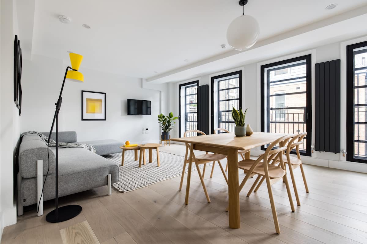 A modern living room with a grey sofa, a wooden dining table with chairs, a yellow floor lamp, and large windows with black shutters.