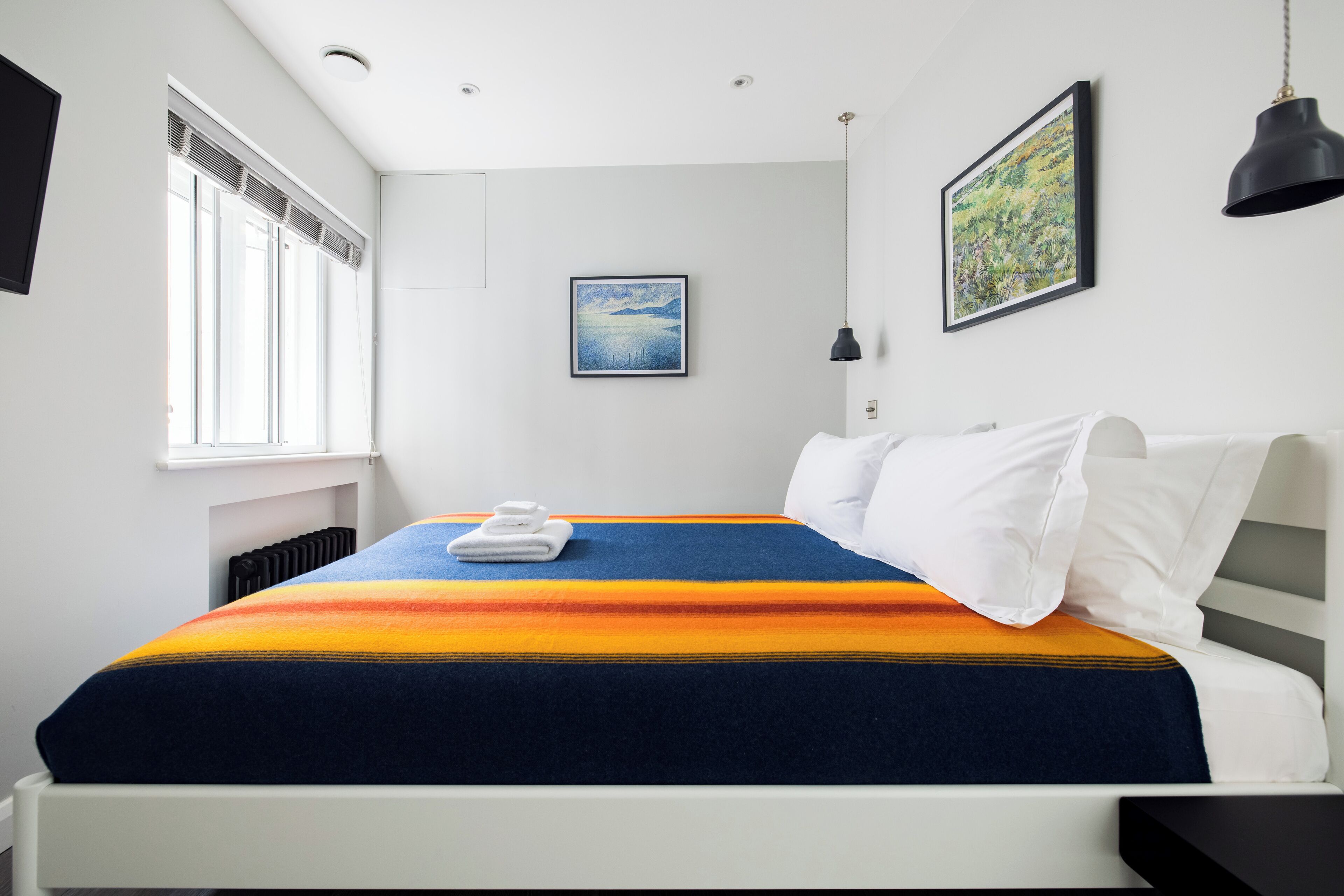 A modern hotel room with a neatly made bed, a window with blinds, and a framed picture on the wall.