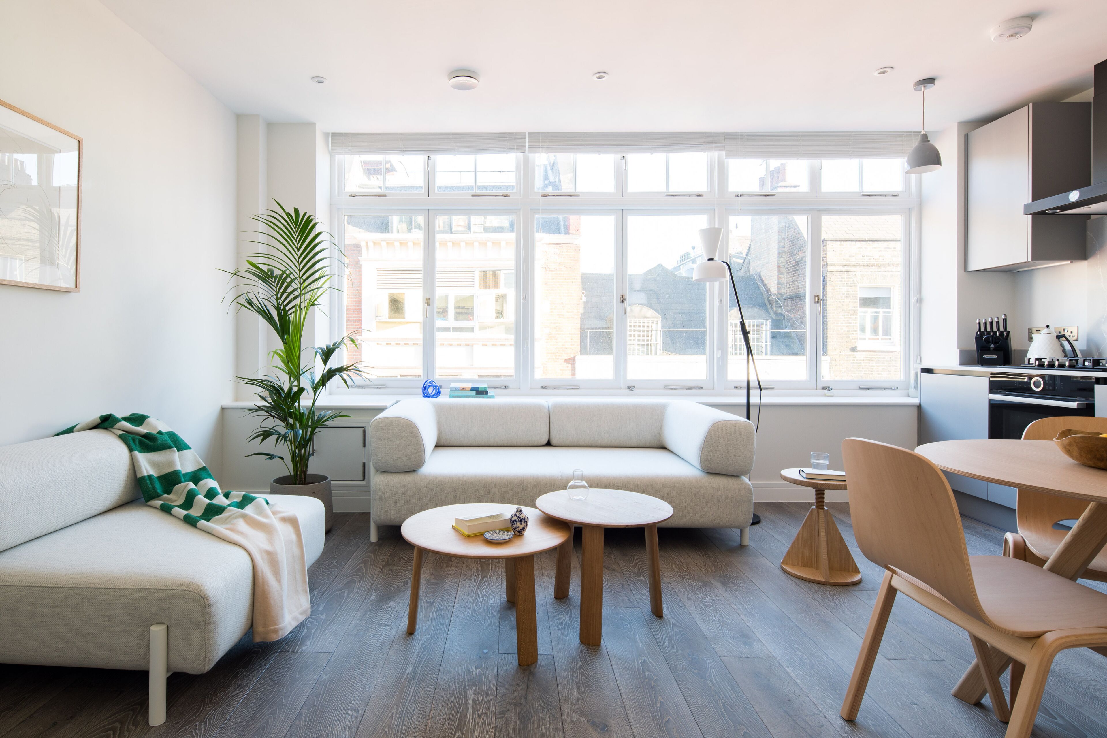 A modern living room with a sofa, two round wooden tables, a dining table with chairs, a potted plant, and a view of buildings through large windows.