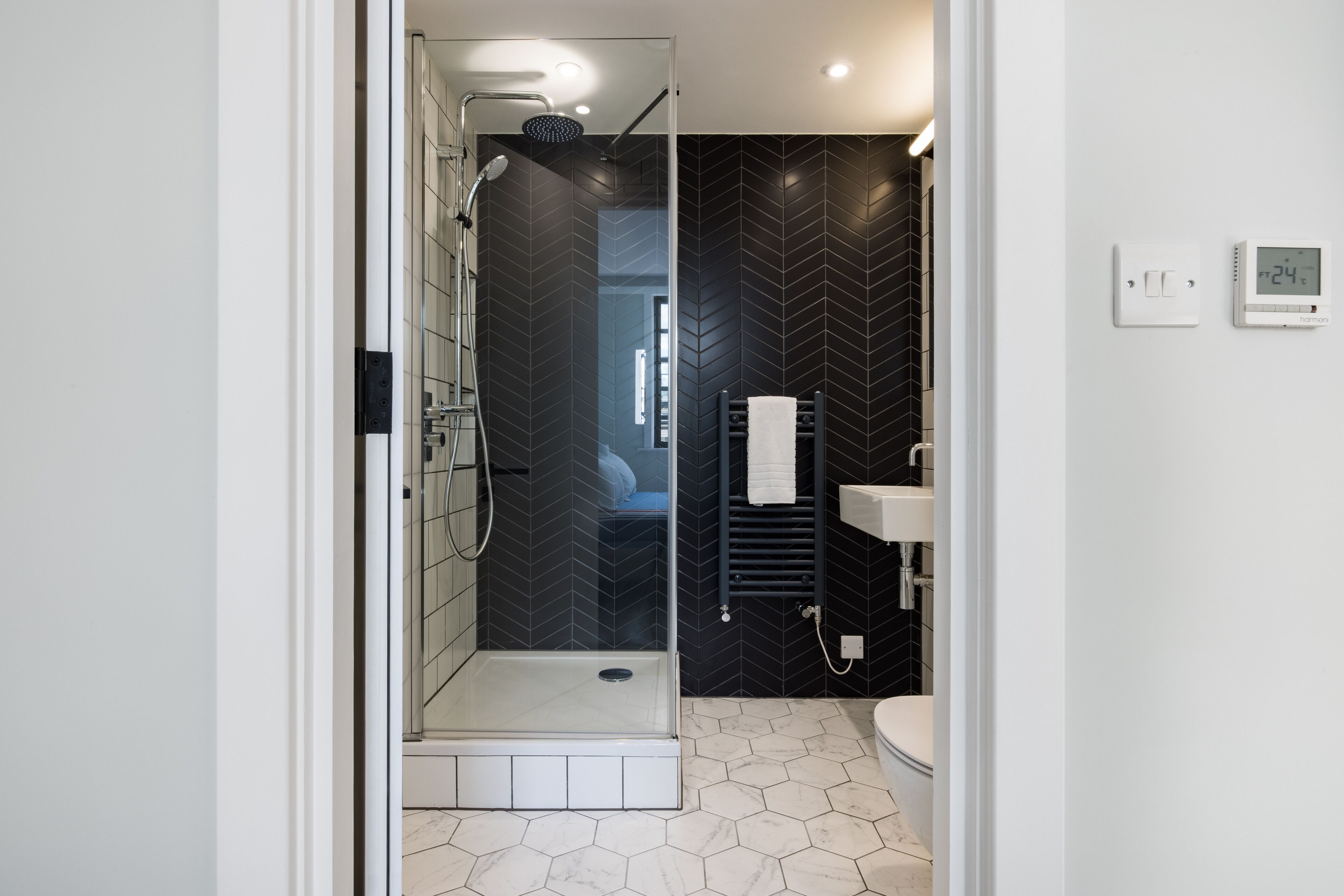 A modern bathroom with a glass-enclosed shower, a black herringbone-patterned wall, a white sink, and a digital thermostat.