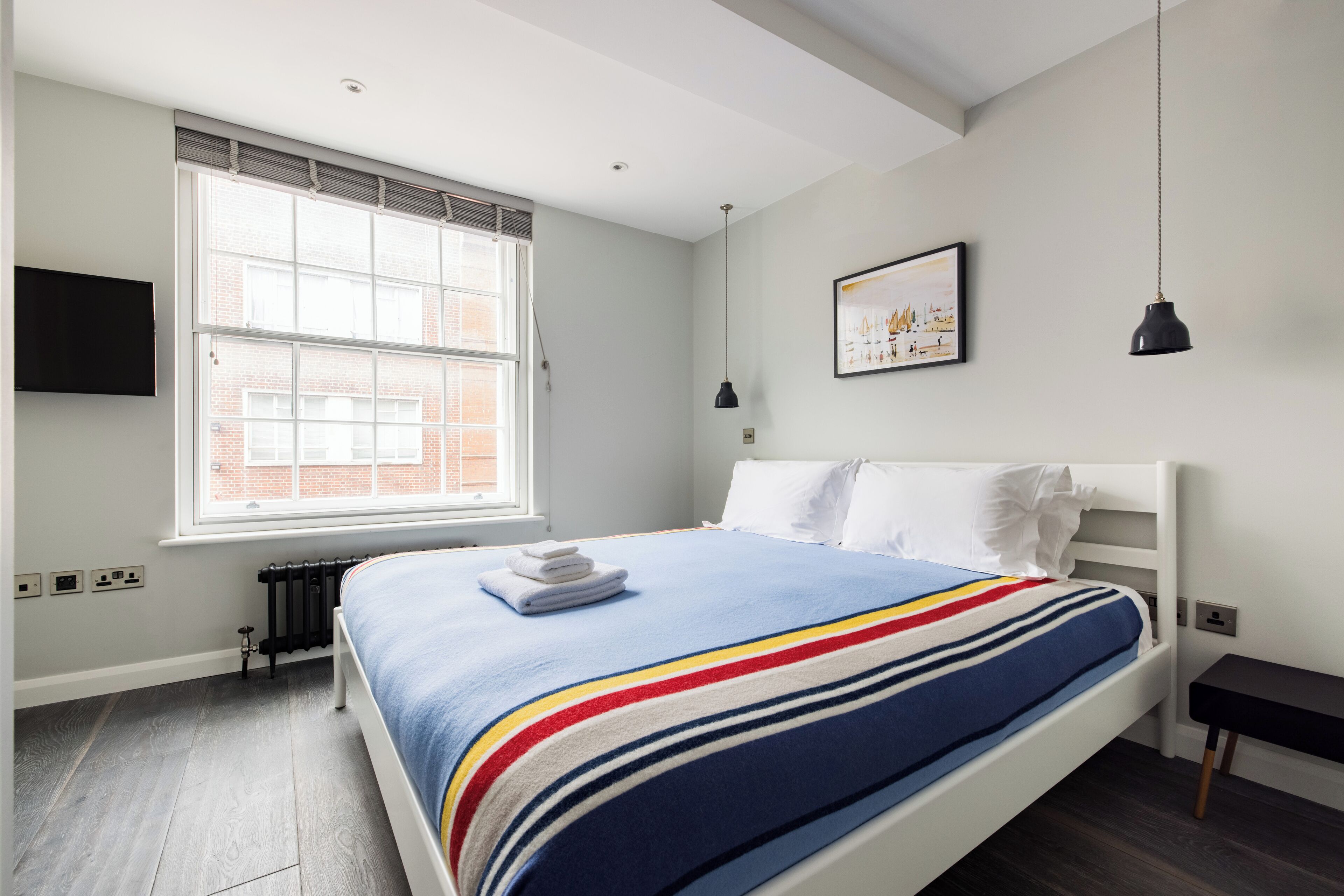 A bedroom with a large window, a bed with a striped blanket, a black desk, and a wall-mounted TV.