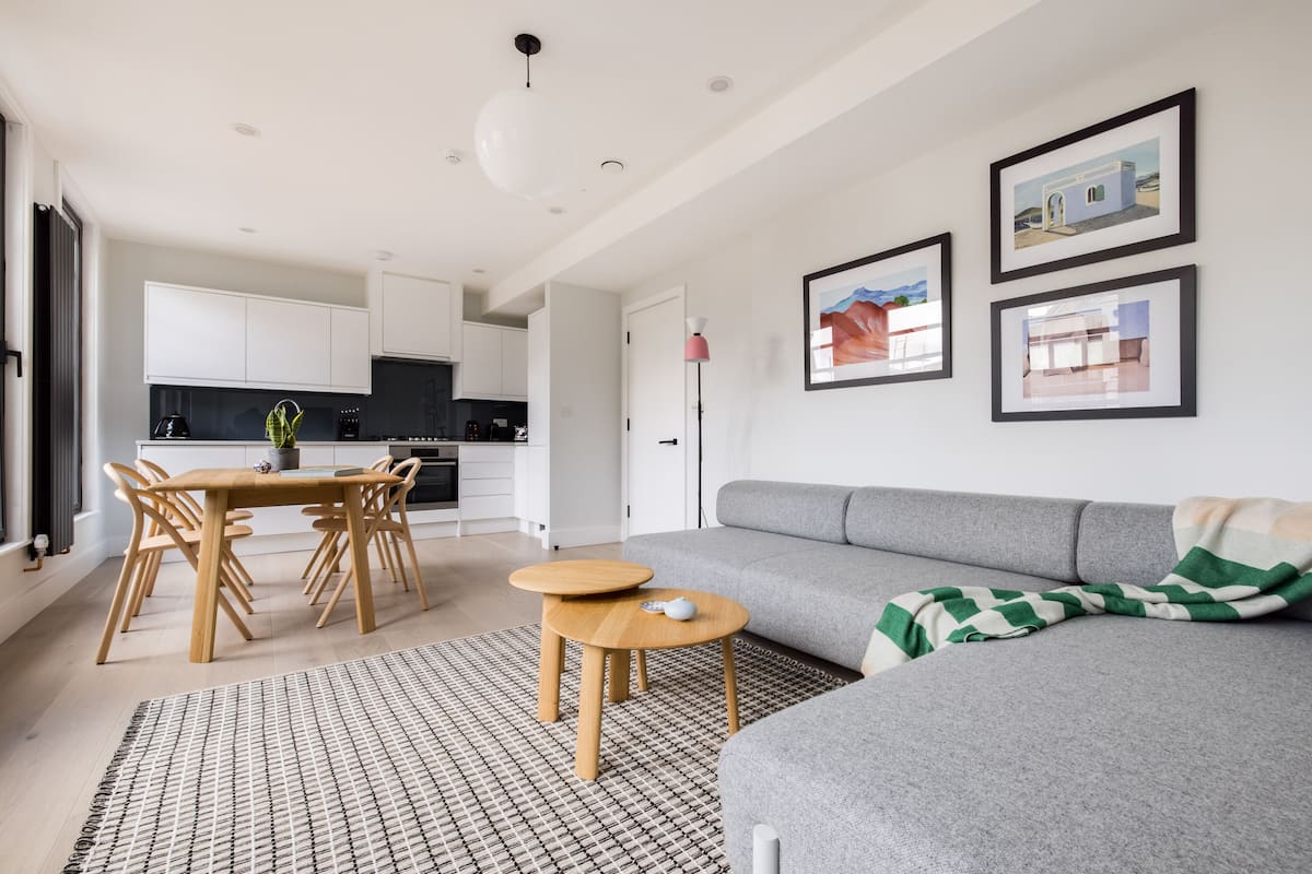 A modern living room with a grey sofa, wooden dining table, and chairs. There are framed pictures on the wall and a small round table with a lamp.