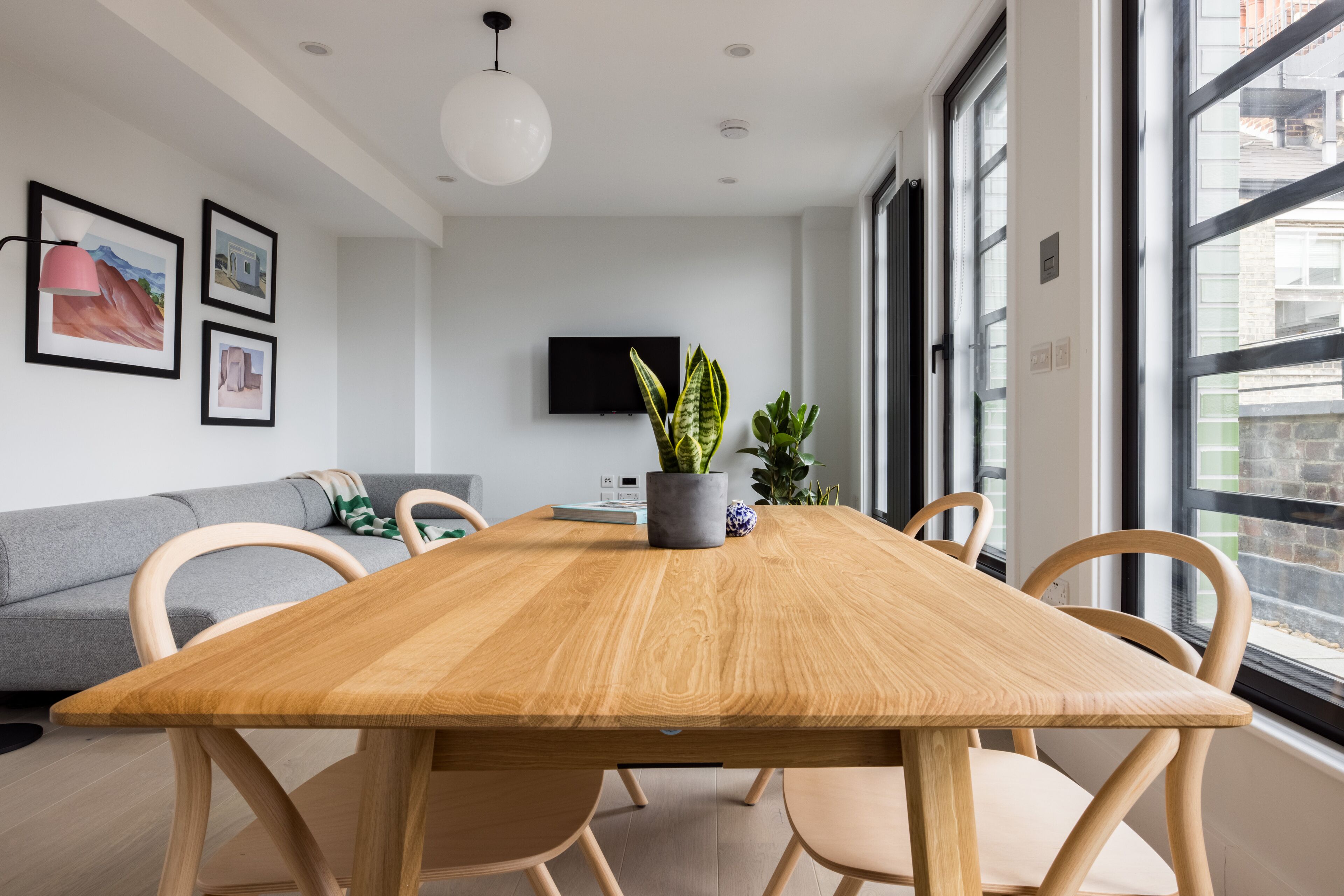 A modern living room with a wooden dining table, grey sofa, and large windows.